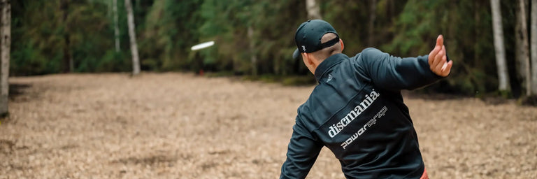 Disc golfer med Discmania logo på ryggen der kaster sin driver hen ad en fairway i en skov